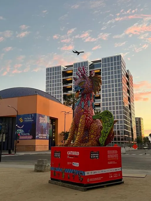 An airplane flies over The Tech Interactive at sunset. CATBIRD, a beautiful Alebrije statue is in the foreground in La Plaza de Cesar Chavez