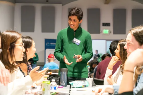 A student in a green sweater with dark hair explains a card game to a group of adults.